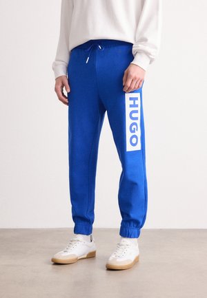 Person iført lyseblå joggers med "HUGO" trykt vertikalt på venstre ben, kombineret med hvide sneakers og en hvid sweatshirt.