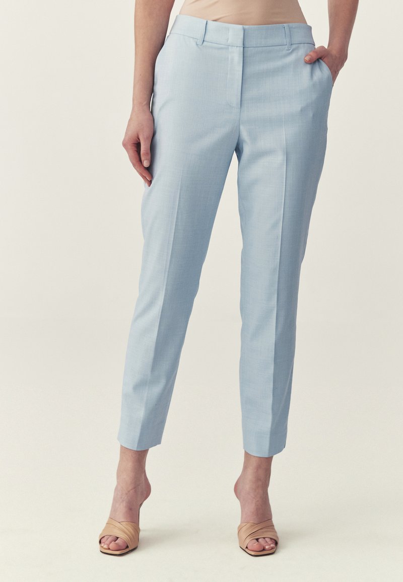 Femme portant un pantalon court bleu clair ajusté avec un pli avant, des sandales à talons beiges, debout avec une main dans la poche contre un fond uni.