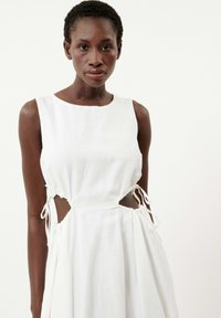 Robe en lin blanc avec un col rond, des découpes sur les côtés et des liens ajustables à la taille. Texture lisse avec une silhouette fluide.