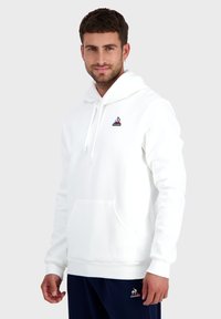Hoodie blanc en tissu doux, avec une poche kangourou, une capuche réglable avec cordon et un petit logo sur la poitrine.
