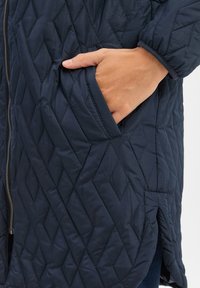 Gesteppten marineblauen Mantel mit einem Reißverschluss vorne und seitlichen Taschen. Der Stoff hat eine glatte Textur mit Rautenmuster und ist mittellang geschnitten.