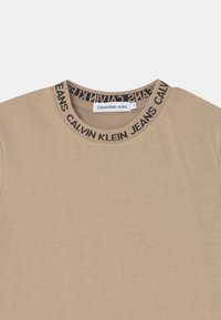 Béžové bavlněné tričko s kulatým výstřihem. Límec má černě opakování loga "Calvin Klein Jeans". Štítek je viditelný uvnitř.