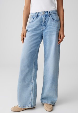 Personne portant un jean large bleu clair avec poches avant, un haut blanc sans manches et des mocassins en daim beige sur un fond uni.