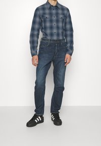 Marinblå rutigt skjorta med knappar och två bröstfickor, parat med mörkblå jeans och svarta skor med vita ränder. Klassisk passform, blandning av bomull.