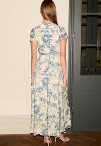 Femme debout à l'intérieur portant une robe maxi crème avec un imprimé paysager bleu et des manches courtes, assortie à des sandales à talons blanches.