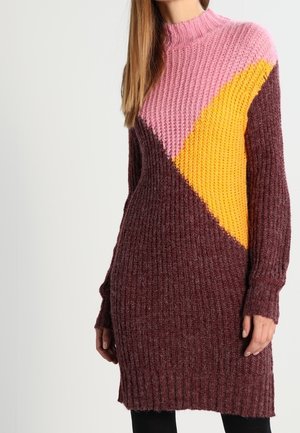 Femme portant une longue robe-pull en tricot avec des blocs de couleur rose, jaune et bordeaux, debout devant un fond clair uni.