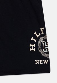 Camiseta de algodón negra con texto gráfico en color crema. El texto incluye "HILFIGER" y "NEW" con un diseño de logo circular.
