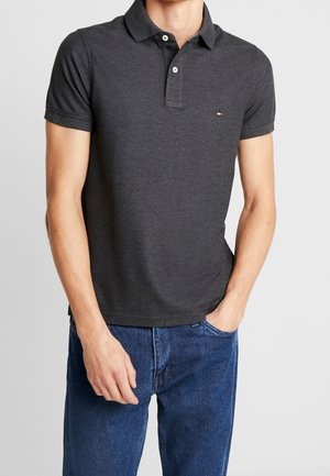 Mann trägt ein dunkelgraues, kurzärmeliges Poloshirt mit zwei Knöpfen und blaue Jeans, eine Hand in der Tasche, vor hellem, einfarbigem Hintergrund.