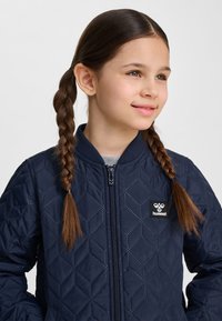 Młoda dziewczyna z brązowymi splecionymi włosami, ubrana w granatową pikowaną kurtkę z zamkiem błyskawicznym z przodu oraz naszywką z logo Hummel.