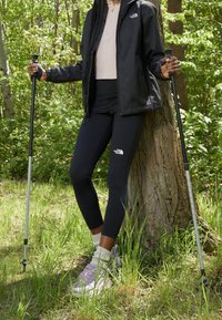 Czarna kurtka z kapturem, jasnoróżowa bluzka, czarne legginsy i szaro-fioletowe buty trekkingowe. Trzymając kije trekkingowe, stoi przy drzewie w zieleni.