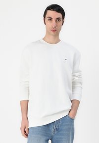 Hvit hettegenser med rund hals, ribbestrikkede mansjetter og kant. Har et lite, flerfarget logo på brystet. Brukt med lys blå jeans.