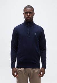 Homme portant un pull à fermeture éclair partielle bleu marine avec le logo "ck" et un pantalon beige, debout devant un fond clair uni.
