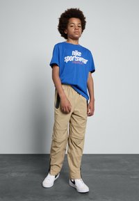 Zils kokvilnas T-krekls ar baltu "Nike Sportswear" logotipu, kombinēts ar brūniem kravas biksēm ar rāvējslēdzēja kabatām un melniem akcentiem, valkāts ar baltām sporta apaviem.