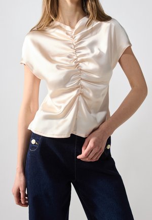 Lyserød satinbluse med rynket frontdesign, korte ærmer og rund halsudskæring. Kombineret med mørke denimbukser med knapdetalje.