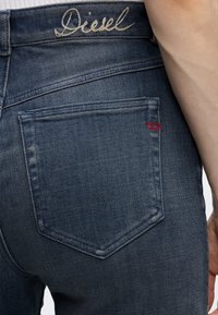 Οπίσθια όψη μπλε τζιν με κεντημένο λογότυπο "Diesel" στη μέση και μια κόκκινη λεπτομέρεια στη γωνία της πίσω τσέπης.