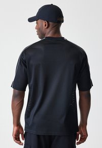 Svart mesh-idrettsjersey med korte ermer, med hvite aksentstriper langs skuldrene og ventilasjonshull for pusteevne.