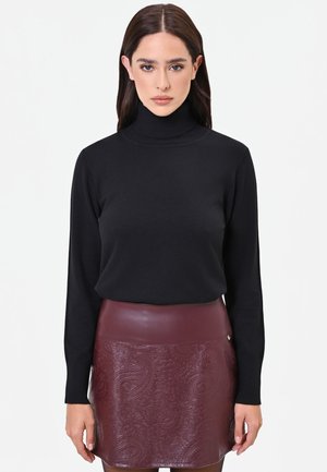 Donna con lunghi capelli scuri che indossa un maglione nero a collo alto e una gonna in pelle bordeaux testurizzata, in piedi davanti a uno sfondo bianco.