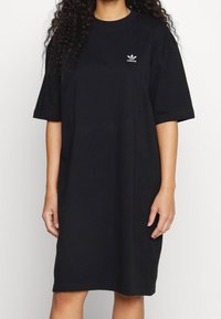 Žena ve volných černých šatech s krátkým rukávem od Adidas, na hrudi s malým bílým logem Adidas.