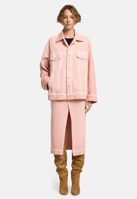 Giacca di denim rosa chiaro con doppie tasche sul petto e chiusura a bottoni, abbinata a una gonna midi rosa coordinata con uno spacco frontale. Stivali marroni fino al ginocchio.