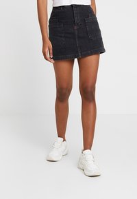 Černá džínová sukně s rozšířeným designem, předními kapsami a švy, doplněná bílými sportovními botami.