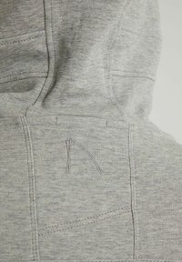 Gros plan du tissu gris clair d’un sweat à capuche montrant des coutures cousues et une lettre brodée "N" au dos, près de la capuche.