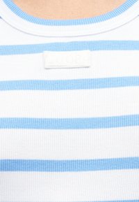 Κοντινό πλάνο ραβδωτού υφάσματος με οριζόντιες λευκές και ανοιχτό μπλε ρίγες και ένα μικρό λευκό ορθογώνιο σημείο κοντά στη λαιμόκοψη.