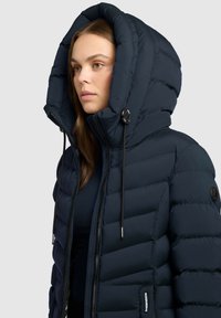 Marine pufferjakke med en stor, quiltet hætte, frontlynlås og snoretræk. Tekstureret stof med synlige sømme og et logo patch.