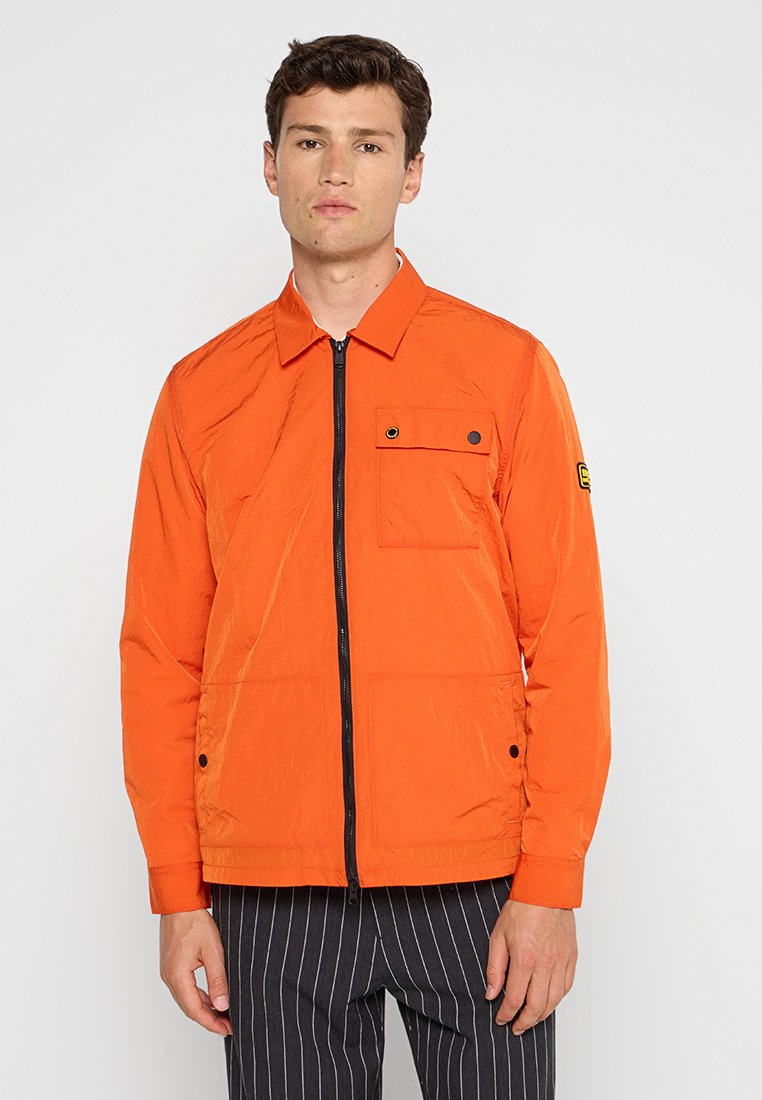 Leichte orangefarbene Jacke mit Reißverschluss, spitzem Kragen, einer Brusttasche und schwarzen Hardware-Akzenten, kombiniert mit gestreiften Hosen.