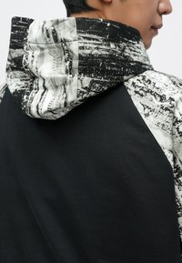 Bluza z kapturem w czarnym kolorze, z teksturowanym kapturem i nakładkami na ramionach w szaro-białym wzorze. Wykonana z mieszanki bawełny.