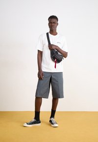 Maglietta bianca con logo "Dickies", pantaloncini grigi, borsa a tracolla nera, calzini neri e sneakers nere, in piedi su un pavimento giallo.