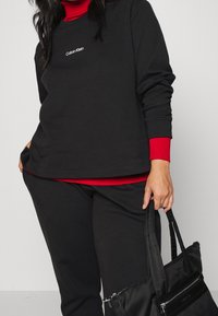 Svart sweatshirt med röda manschetter och en logotyp vid bröstet, ihopparad med matchande svarta byxor och en svart tote-väska med dragkedjedetalj.