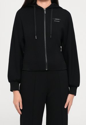 Femme portant un sweat à capuche noir zippé avec cordons et logo Armani Exchange, assorti à un pantalon noir coordonné.