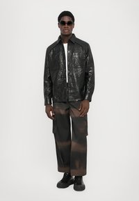 Black crinkled leather jacket with two front pockets and a collared neckline, paired with brown cargo pants featuring a distressed pattern.