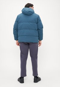 Padded, kaputast rukavac u tirkiznoj boji s suptilnim sjajem, s vodoravnim kviltiranim dijelovima. Uparen s tamnosivim hlačama i crnim cipelama.
