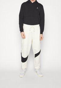 Schwarzer Kapuzenpullover mit einer Vordertasche, kombiniert mit weißen Jogginghosen, die schwarze gebogene Akzente und einen elastischen Bund aufweisen.