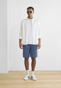 Camisa blanca de botones sobre una camiseta blanca, combinada con pantalones cortos azules y zapatillas blancas con rayas negras. Unas gafas de sol elegantes completan el look.