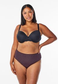 Žena so stredným tónom pleti, oblečená v čiernej podprsenke a hnedých nohavičkách s vysokým pásom s malými ružovými srdcami, stojí s jednou rukou na bok.