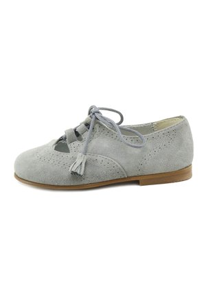 Zapato plano de ante gris claro con detalles perforados, cordones delanteros con borlas y suela baja marrón.