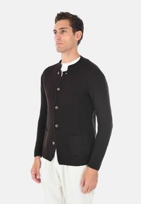 Cardigan marrone lavorato a maglia con scollatura rotonda, grandi bottoni e due tasche frontali. Il tessuto presenta un motivo strutturato, abbinato a pantaloni bianchi.