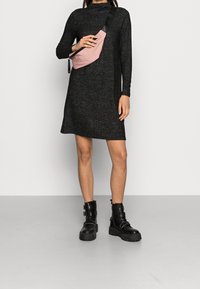 Robe noire à manches longues avec une coupe décontractée. Un sac bandoulière en tissu rose est porté en diagonale sur le torse. Des bottes noires épaisses avec des sangles complètent le look.