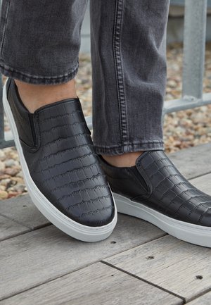Baskets noires à enfiler avec un motif texturé, portées avec un jean gris foncé sur une surface en bois près de gravier et d'une rambarde métallique.