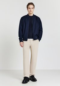 Jeune homme aux cheveux châtains clairs portant une veste bomber bleu marine, un pull sombre, un pantalon beige et des chaussures noires, se tenant contre un fond blanc.