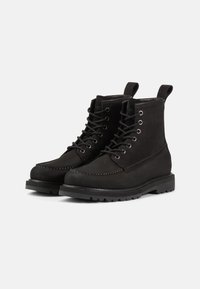 Botas de tobillo de ante negro con ojales de metal, diseño de cordones y suela tipo lug. Presentan un detalle de costura en contraste y una pestaña en la parte trasera.