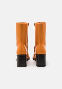 Botas de tobillo de cuero naranja con una textura suave, que cuentan con un cierre de cremallera en la parte trasera y un tacón grueso negro, diseñadas para una silueta ajustada.