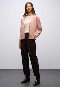 Světle růžový kardigan s texturovanými, vroubkovanými okraji a knoflíky, doplněný krémovým topem a tmavě hnědými kalhotami. Elegantní černé boty do kotníků.