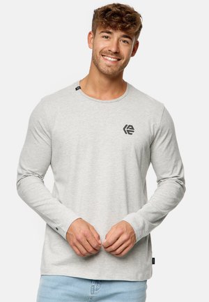 Camisa de manga larga gris hecha de un tejido suave, con un cuello redondeado y un pequeño logo negro en el lado izquierdo del pecho. Diseño sencillo con puños ajustados.