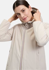 Veste imperméable beige clair avec capuche, dotée d'une fermeture éclair à l'avant bordée de rayures colorées et de deux poches latérales. Texture de tissu lisse.