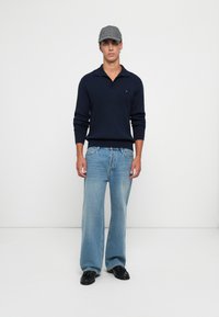 Jonge man in een grijze pet, marineblauwe trui, lichtblauwe wijde jeans en zwarte schoenen, staande tegen een effen witte achtergrond.