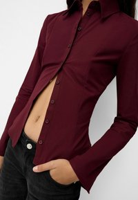 Camicia fitted bordeaux con colletto a punta, apertura con bottoni e maniche lunghe; tessuto liscio con una leggera lucentezza.