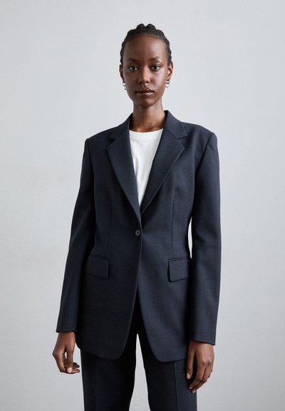 Mørkeblå blazer med strukturerede skuldre, enkelt knaplukning og to frontlommer. Båret over en hvid skjorte med en glat tekstur.